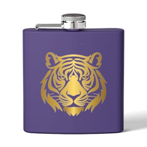 Puede incluir: Un petaca morada de acero inoxidable con un diseño de cabeza de tigre dorada. La petaca tiene una tapa con bisagras plateada y forma rectangular. La cabeza del tigre está centrada.