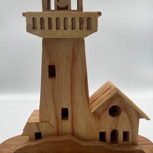 May include: A wooden lighthouse model with a detailed design. The lighthouse features a tower, a lantern room, and an attached house, all crafted from light-colored wood. The model sits on a round wooden base, showcasing the natural wood grain.
