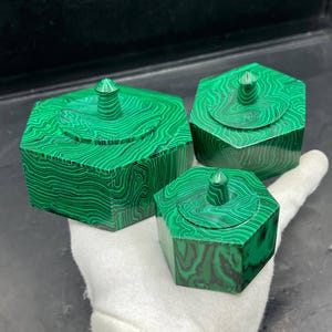 Può includere: Tre scatole esagonali con coperchi, realizzate in pietra di malachite verde. Le scatole sono di diverse dimensioni e presentano un motivo a vortice e a fasce in verde e nero. Ogni scatola ha un piccolo pomello arrotondato sul coperchio.