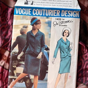 May include: A Vogue Couturier Design sewing pattern envelope, number 1423 by Jo Mattli of London. The front features a photograph of a person in a dark textured suit and a blue hat, alongside a fashion illustration of the same suit in light blue.