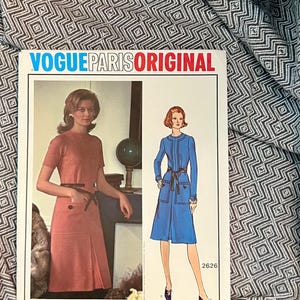 Peut inclure: Une pochette de patron de couture Vogue Paris Original 2626 par Molyneux. Le recto présente une photographie d'une personne portant une robe corail à manches courtes avec une ceinture noire et une illustration d'une robe bleue à manches longues.