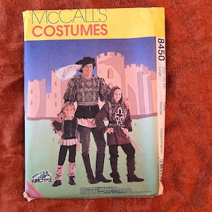 Puede incluir: Patrón de costura McCall's Costumes número 8450 para un disfraz medieval de talla grande (42-44). El patrón incluye instrucciones para hacer una túnica, pantalones y botas. El patrón está ilustrado con tres personas que llevan los disfraces.