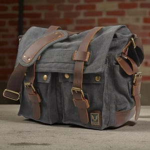 May include: Grey canvas messenger bag with brown leather detailing. Features an adjustable shoulder strap, multiple pockets, and brass-coloured buckles. The bag is rectangular with a flap closure.