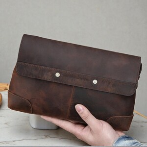 Könnte beinhalten: Eine braune Leder-Clutch mit Klappe und zwei silbernen Druckknöpfen. Die Clutch hat eine Used-Optik und eine rechteckige Form. Der Text "Concord Genuine Leather Co." ist auf der Klappe sichtbar.