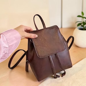 Puede incluir: Una mochila de cuero marrón oscuro con un asa superior, una solapa con cierre magnético y dos correas delgadas para los hombros. El compartimento principal está asegurado con un cierre de cordón y el exterior tiene un acabado mate.