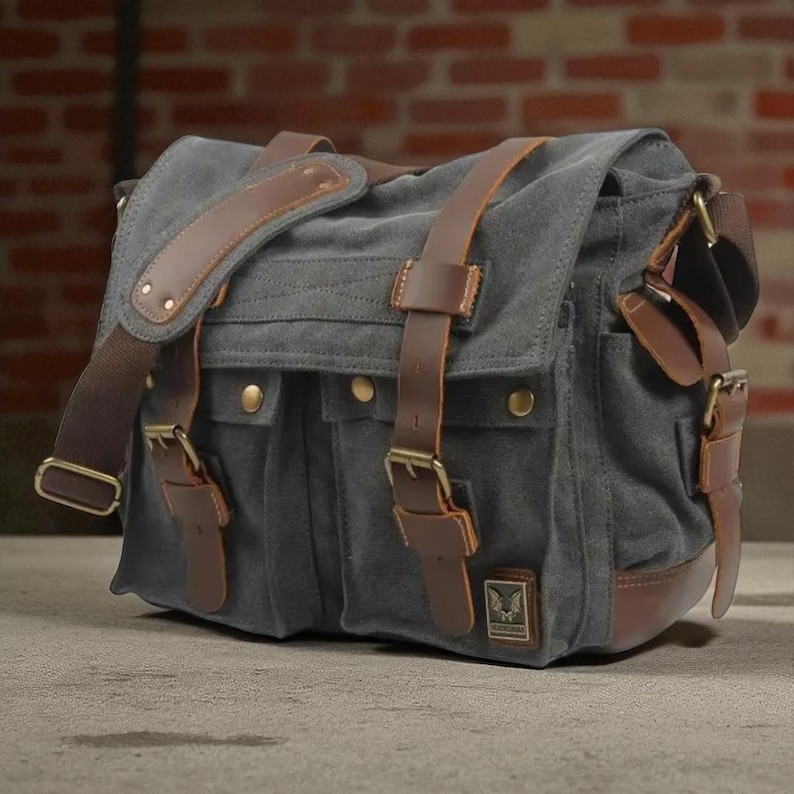 May include: Grey canvas messenger bag with brown leather detailing. Features an adjustable shoulder strap, multiple pockets, and brass-coloured buckles. The bag is rectangular with a flap closure.