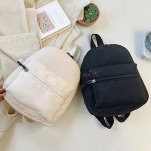 May include: Two small backpacks, one in off-white and one in black, each featuring a main compartment and a front zippered pocket with silver-toned hardware. The bags have adjustable shoulder straps and a top handle for carrying.