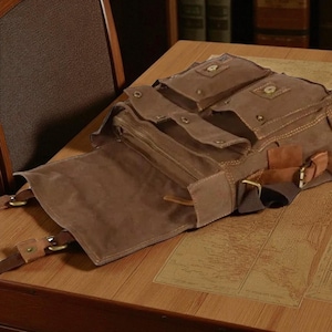 Op de afbeelding: Een bruine canvas rugzak met meerdere zakken en compartimenten. De tas heeft leren accenten, metalen gespen en een hoofdvak met ritssluiting. De tas is open, waardoor de binnenkant zichtbaar is, en ligt op een houten oppervlak.
