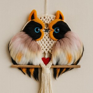 Op de afbeelding: Een handgemaakte wanddecoratie met twee uilfiguren met zwarte, oranje en roze veren, blauwe ogen en een rood hart. De uilen zitten op een houten deuvel, verbonden door macramé en een crèmekleurige kwast.