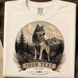 Może przedstawiać: Kremowy t-shirt z okrągłą grafiką przedstawiającą wilka stojącego na skale w górskim lesie. Pod wilkiem znajduje się baner z napisem "YOUR TEXT", "EST. 2026" oraz "YOUR LOCATION" z małą ikoną góry pomiędzy liniami tekstu.