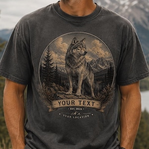 Może przedstawiać: Ciemnoszary t-shirt z krótkim rękawem z okrągłą grafiką przedstawiającą wilka stojącego w lesie na tle gór. Projekt zawiera baner z napisem YOUR TEXT, EST. 2026 oraz YOUR LOCATION wydrukowanym pod wizerunkiem wilka.