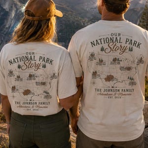 May include: Two cream-colored t-shirts featuring a graphic of the United States map with national park locations, the text "OUR NATIONAL PARK Story," "THE JOHNSON FAMILY," "Adventures & Memories," and "EST. 2025" printed on the back of each shirt.