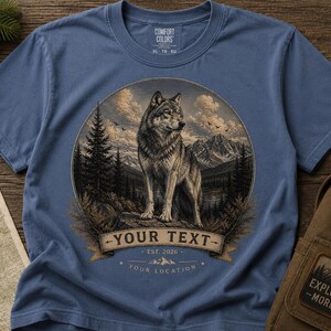 May include: A blue t-shirt featuring a circular graphic of a wolf standing in a forest with mountains in the background. The design includes a banner with the text YOUR TEXT, EST. 2026, and YOUR LOCATION. The shirt has a Comfort Colors label.