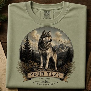 May include: A sage green crew neck t-shirt featuring a circular graphic of a wolf standing on a rock in a mountain forest landscape. Below the wolf is a banner with the text YOUR TEXT, EST. 2026, and YOUR LOCATION. The shirt has a Comfort Colors label.