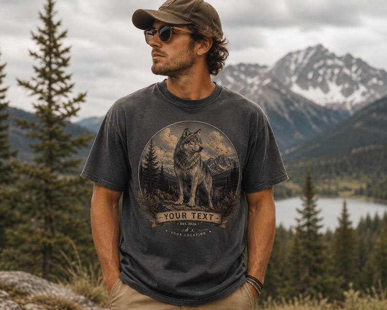 Może przedstawiać: Ciemnoszary t-shirt z krótkim rękawem z okrągłą grafiką przedstawiającą wilka stojącego w lesie na tle gór. Projekt zawiera baner z napisem YOUR TEXT, EST. 2026 oraz YOUR LOCATION wydrukowanym pod wizerunkiem wilka.