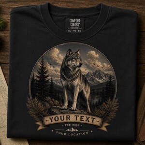 May include: A black t-shirt featuring a circular graphic of a wolf standing on a rock in a mountain landscape with pine trees and clouds. Below the wolf is a banner with the text YOUR TEXT, EST. 2026, and YOUR LOCATION. The shirt has a Comfort Colors label.