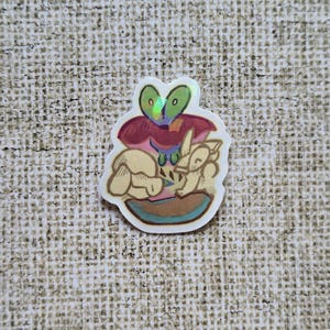 Puede incluir: Una pegatina con una ilustración caprichosa de una criatura parecida a una rana con ojos verdes y una cabeza roja y morada. La criatura descansa sobre una base marrón y azul, con dos figuras más pequeñas de color claro. La pegatina tiene un borde blanco.