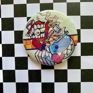 Puede incluir: Un botón redondo con una ilustración de dibujos animados de dos personajes con aspecto de monstruo. Uno es rojo con la boca muy abierta y el otro azul con una lengua larga. El botón tiene un fondo azul claro con un borde colorido.