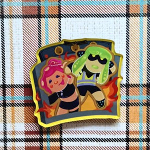 May include: A colorful pin featuring two cartoon characters in front of a gray oven with flames. One character has pink hair and the other has green hair. The pin has a yellow border and is set against a plaid background.