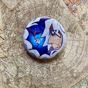 May include: A round button featuring a stylized anime character profile with white hair and a blue and white background. The button has a central image of a red and white creature. The button is on a vintage map.