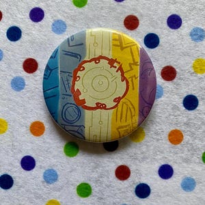 May include: A round button with a colorful design. The button features vertical stripes in blue, yellow, and purple, with a central design in red and cream. The button is likely a pin or badge.