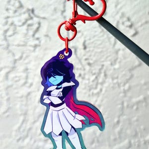 May include: A cartoon keychain featuring a character with blue skin, black hair, and a white outfit with a red cape. The keychain has a red heart-shaped clasp and a small ring. The background is a textured white surface.