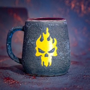 Puede incluir: Una taza de cerámica con un exterior gris carbón texturizado y un interior liso de color rojo intenso. La parte frontal presenta un gráfico amarillo brillante de una calavera estilizada con llamas sobre la superficie rugosa.