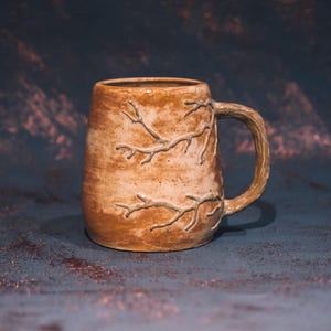 Może przedstawiać: Ręcznie wykonany ceramiczny kubek o teksturowanym, ziemistym brązowym wykończeniu. Kubek posiada wzór gałęzi drzewa wyrzeźbiony na powierzchni i pasujący uchwyt w kształcie gałęzi. Kubek ma około 10 cm wysokości.