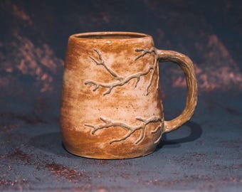 May include: A handmade ceramic mug with a textured, earthy brown finish. The mug features a tree branch design carved into the surface and a matching branch-shaped handle. The mug is approximately 4 inches tall.