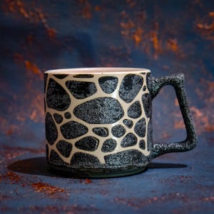 Puede incluir: Una taza de cerámica con un patrón texturizado de piedra en negro y gris oscuro, con líneas suaves de color crema que forman una cuadrícula inspirada en una jirafa. La taza tiene un asa angular gruesa con una superficie oscura y porosa.