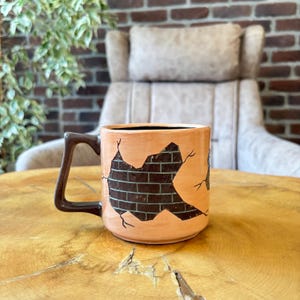 Op de afbeelding: Een keramische koffiemok met een bruin handvat en een uniek ontwerp. De mok heeft een perzikroze buitenkant met een bruin baksteenpatroon, waardoor een visueel effect ontstaat van een muur die doorbreekt. De binnenkant is zwart.