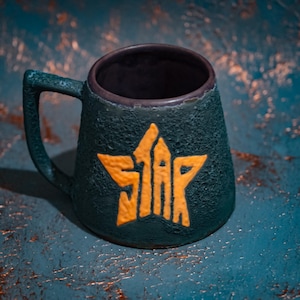 Puede incluir: Una taza de cerámica verde oscuro con una superficie rugosa y texturizada y un asa. La parte frontal de la taza presenta un emblema naranja en relieve con forma de estrella, con la palabra STAR integrada en el diseño con una fuente llamativa.