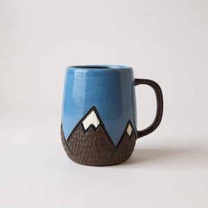 May include: A ceramic mug with a blue sky design and a mountain range. The mountains are brown with white snow caps. The handle is brown. The mug is approximately 4 inches tall.