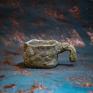 May include: A handmade ceramic mug with a textured, cracked stone effect. The grey mug has yellow-green detailing in the cracks and a curved handle. The mug is designed for drinks and has a rustic, artistic appearance. The mug's dimensions are in centimetres.