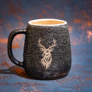 Peut inclure: Un mug en céramique avec une surface extérieure gris foncé, fortement texturée et granuleuse, et un intérieur lisse de couleur crème clair. L'avant du mug présente une illustration au trait claire d'une tête de cerf avec des bois.