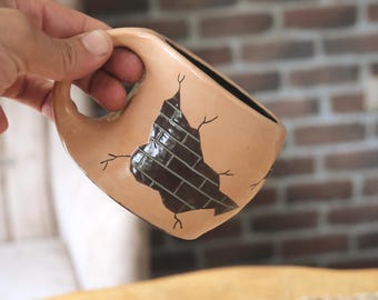 May include: A beige ceramic mug with a unique design. The mug features a dark brown brick pattern within a cutout shape, with black lines radiating outwards. The interior of the mug is black, and it has a curved handle.