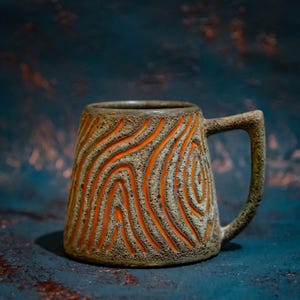 Könnte beinhalten: Eine handgefertigte Keramiktasse mit einem einzigartigen Holzmaserungsdesign. Die Tasse hat eine strukturierte, erdfarbene braune Außenseite mit orangefarbenen Rillen und einen Griff. Die Tasse ist für Heißgetränke konzipiert.