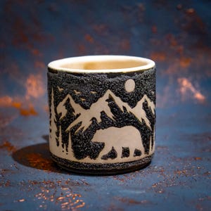 Puede incluir: Una taza cilíndrica de cerámica con un exterior negro texturizado que presenta un diseño tallado de un oso caminando por un bosque hacia picos montañosos bajo una luna llena. El interior de la taza tiene un acabado de cerámica color crema.