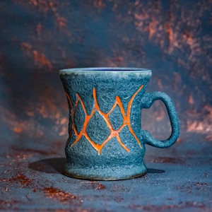 May include: A textured, teal-colored ceramic mug with an orange geometric design. The mug has a curved handle and a slightly flared rim. The background is a mottled mix of teal and orange.
