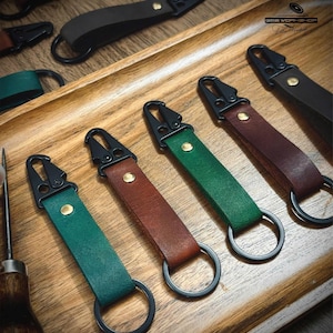 May include: A selection of leather keychains in shades of green, brown, and black. Each keychain has a black metal clasp and a matching ring. The keychains are presented on a wooden tray, highlighting their design.
