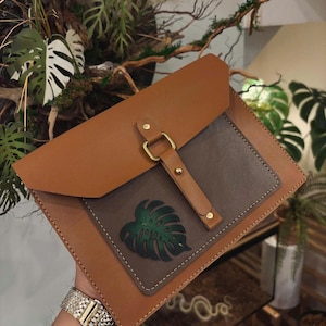 May include: A tan envelope-style bag featuring a dark gray rectangular front pocket with a green monstera leaf applique. The bag has a tan flap closure secured by a vertical strap with a gold-toned metal buckle and two gold-toned rivets.