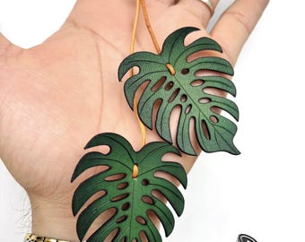 Puede incluir: Dos adornos de cuero verde con forma de hoja de Monstera, suspendidos por un cordón de cuero marrón claro. Las hojas tienen intrincados detalles recortados. El texto "GBB.WORKSHOP Leather Handmade" es visible.