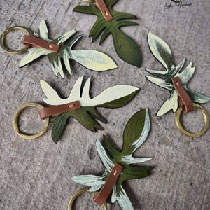 May include: Keychains in the shape of leaves, featuring a combination of green and white paint. Each keychain has a brown leather strap and a gold-colored ring. The keychains are arranged on a textured, gray surface. The text "GSB.WORKSHOP Leather Handmade" is visible.