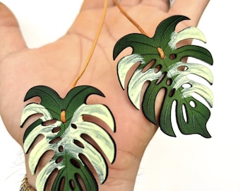 Puede incluir: Dos colgantes de cuero con forma de hoja de monstera, verdes y blancos, suspendidos de cordones color beige. Los colgantes se sostienen en una mano, con un reloj dorado y plateado en la muñeca. El texto "GBB.WORKSHOP Leather Handmade" es visible.