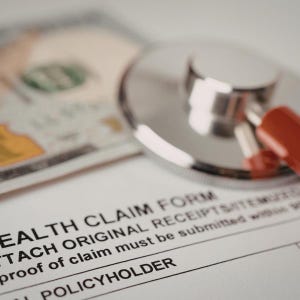 May include: A close-up shot of a health claim form with a stethoscope and a US $100 bill. The form text includes "ATTACH ORIGINAL RECEIPTS" and "POLICYHOLDER". The stethoscope is silver with a red component.
