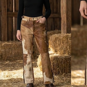 Könnte beinhalten: Hose aus braun-weißem Kuhfell mit ausgestelltem Bein. Die Hose hat eine hohe Taille und eine dunkelbraune Naht an der Seite. Das Model trägt einen schwarzen Rollkragenpullover. Der Hintergrund zeigt Heuballen und Holzbalken.