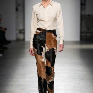 Könnte beinhalten: Ein Model trägt ein cremefarbenes Hemd mit Knöpfen und eine Hose mit Kuhfellmuster. Die Hose hat ein Patchwork-Design mit schwarzen, braunen und weißen Abschnitten. Das Outfit wird durch einen schwarzen Gürtel und Schuhe ergänzt. Der Hintergrund ist ein neutrales Grau.