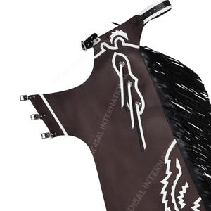 Könnte beinhalten: Braune Lederchaps mit weißen Ziernähten und schwarzem Fransenbesatz. Die Chaps haben verstellbare Schnallen und einen weißen Ledergürtel. Das Design beinhaltet ein weißes Dekorationsmuster.