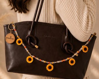 Könnte beinhalten: Eine dunkelbraune Ledertasche mit zwei Schulterriemen und einer dekorativen Perlenkette mit kleinen gelben Sonnenblumenanhängern. Ein rundes, hellbraunes Lederetikett mit einem Logo ist seitlich an der Tasche nahe der Riemenbefestigung angebracht.