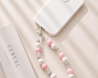 Könnte beinhalten: Ein weißes Smartphone mit einer Perlen-Handschlaufe, bestehend aus abwechselnd rosa, weißen und klaren facettierten Perlen sowie kleinen silberfarbenen, texturierten Kugeln. Die Schlaufe ist mit einem silbernen Metallkarabiner am Telefon befestigt.